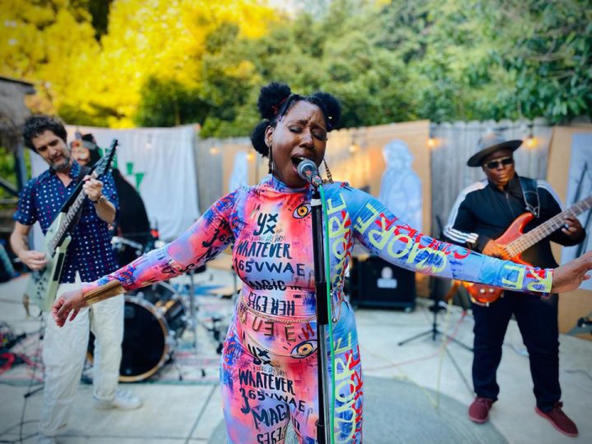 Cardboard People performing in the backyard — singer mid-note with guitarist and bassist behind her
